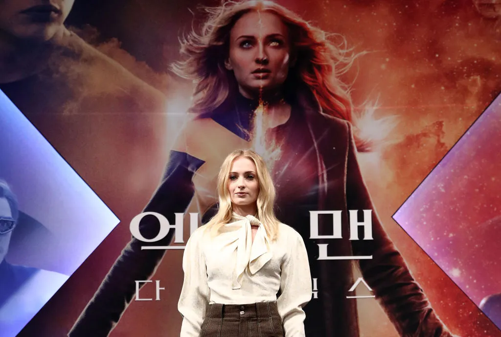 Sophie Turner posing in front of a poster of her playing Jean Grey the mutant 