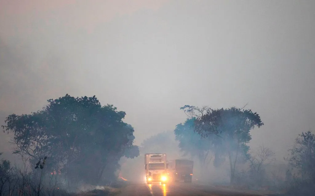 Trucks blare their headlights while driving down a densely smokey road.