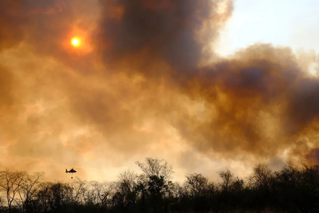 Heavy smoke blocks the sun as a silhouetted helicopter carries water towards the flames.