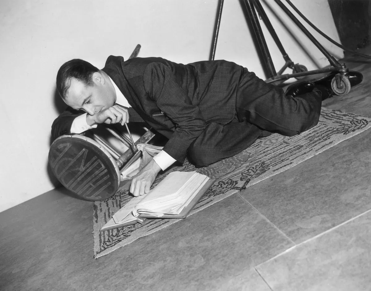 American film director Richard Thorpe reclines on the carpet, reading a script, while resting his chin on his hand.