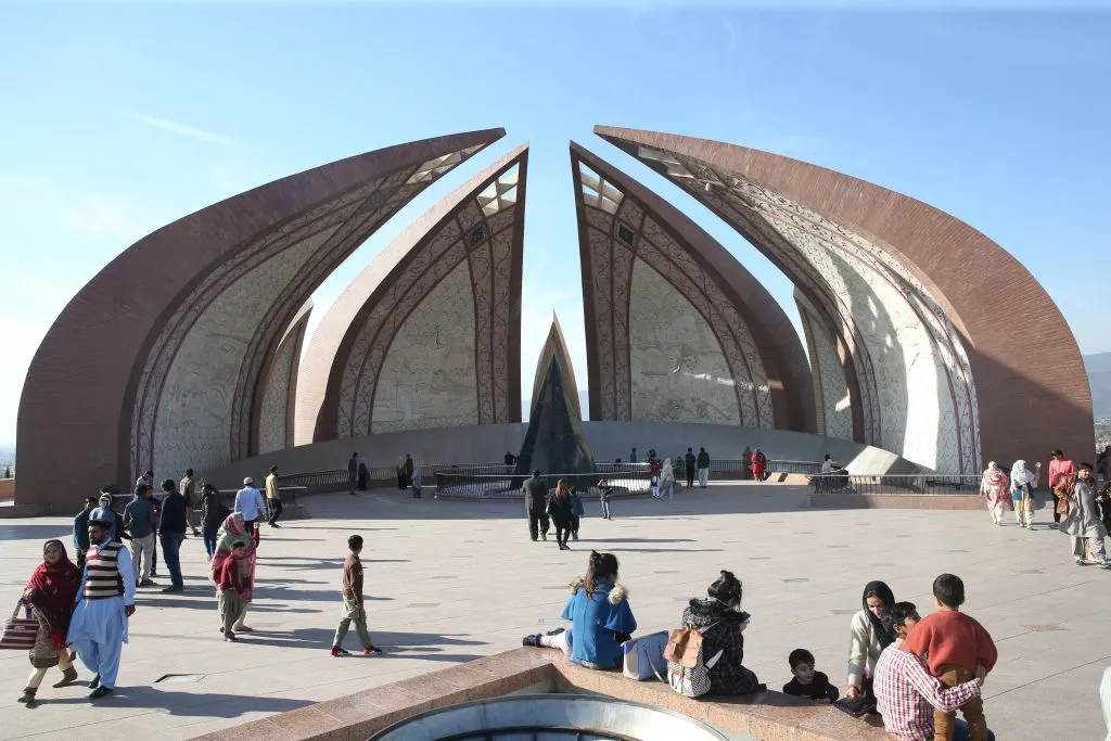 People tour a monument in Islamabad that consists of four pointed arches curved towards one another