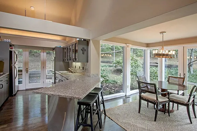 A long kitchen looks on to a breakfast nook surrounded by glass windows.