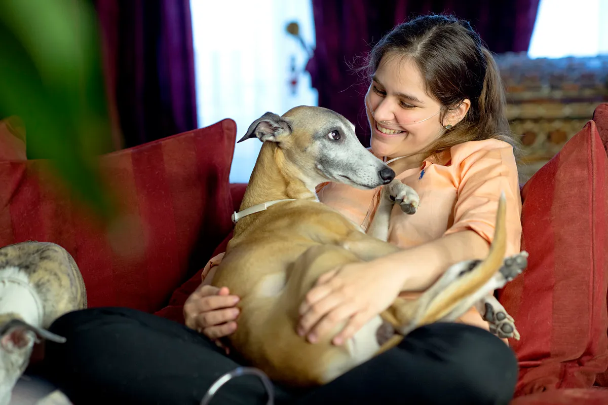 Girl with nasal cannula sits on a sofa and holds her Greyhound in her arms.