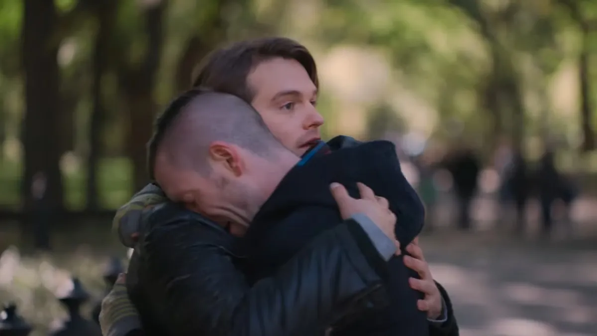 Two men hug looking distraught in a park.