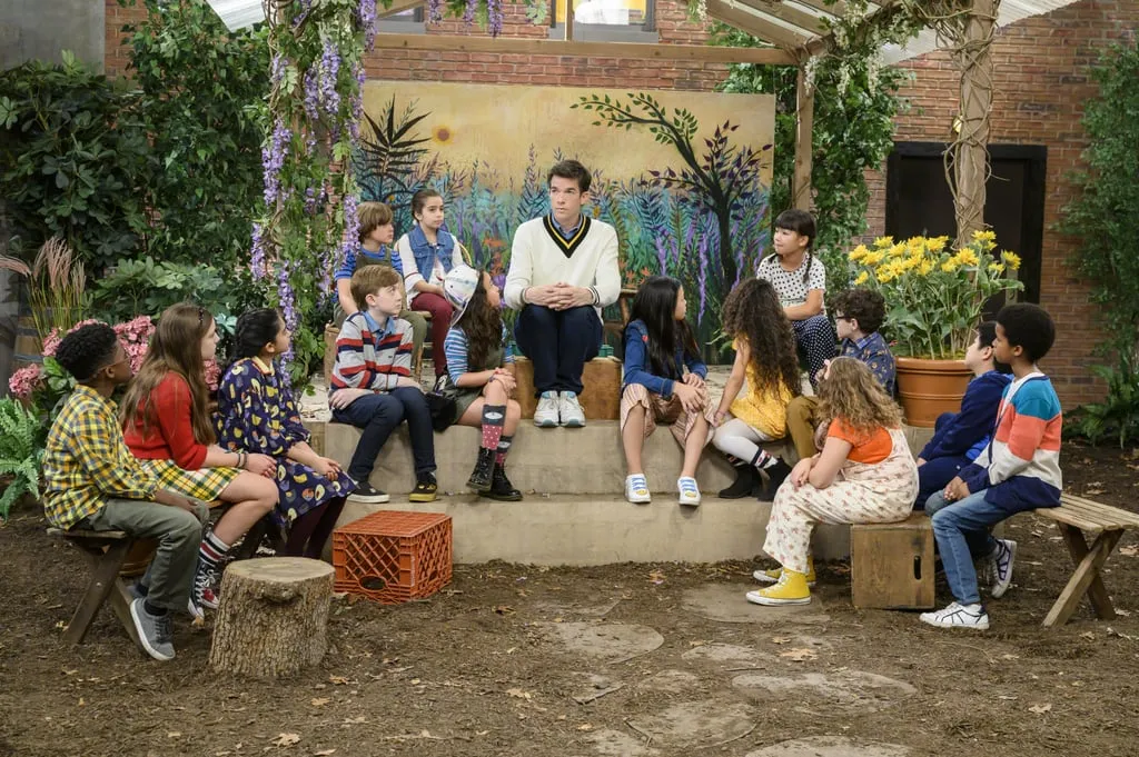 John Mulaney sits in a courtyard amongst children.