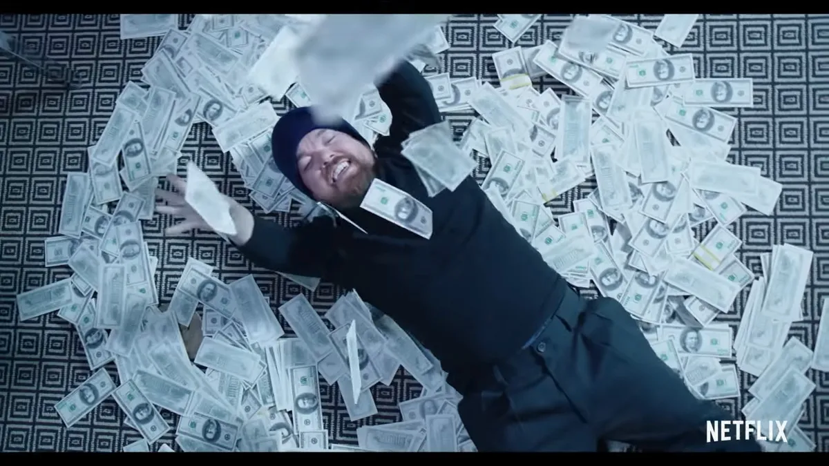A man in all black clothing rolls around in $100 bills.