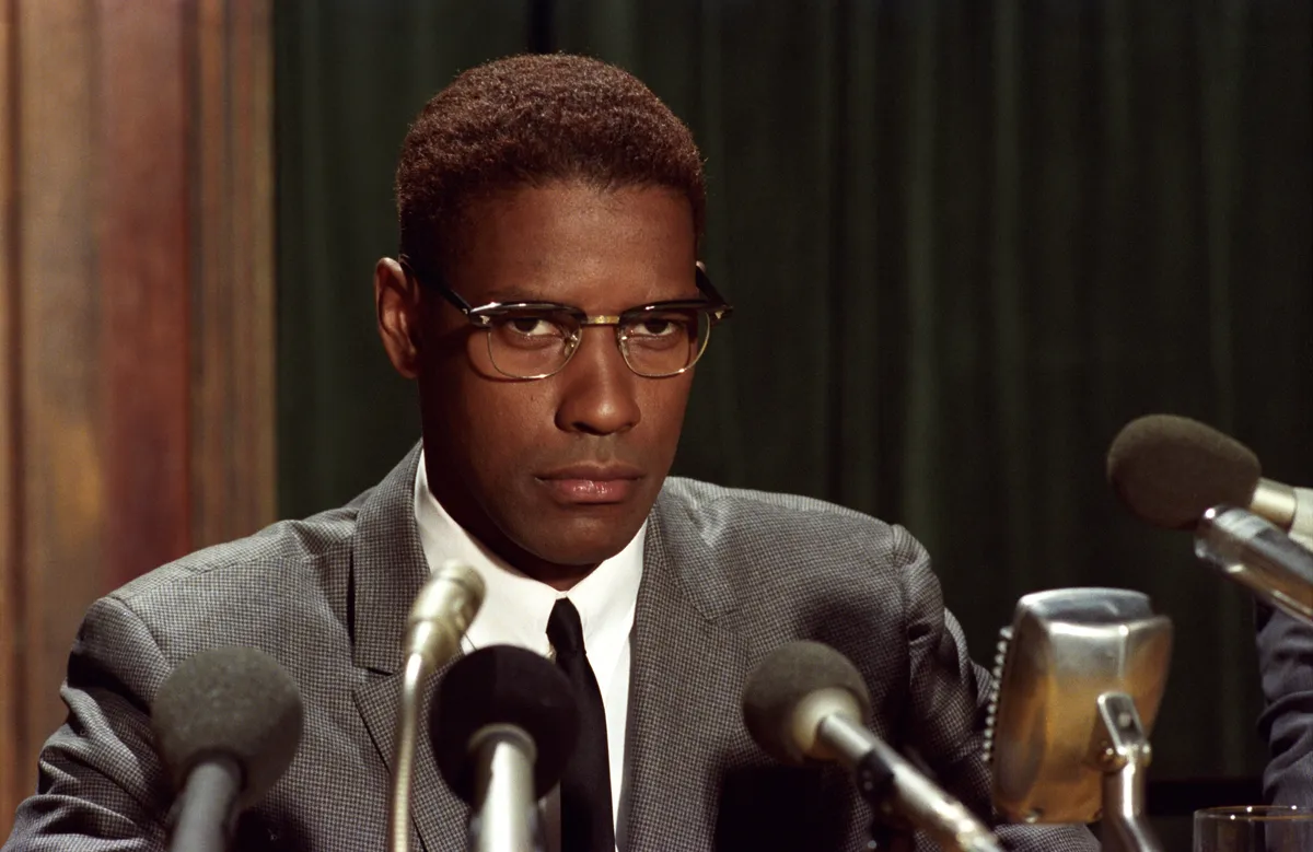 Denzel Washington wears glasses and a serious look while sitting at a press conference.