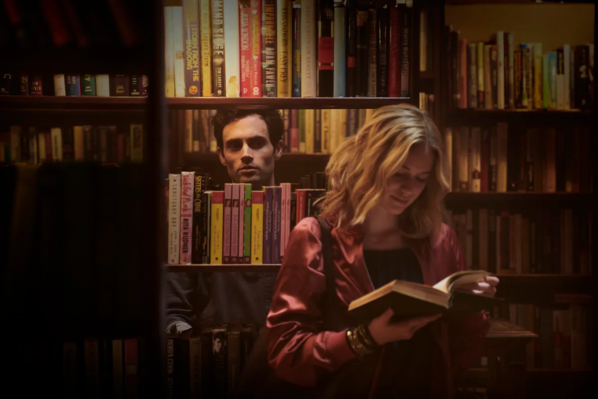 A man looks through a bookshelf at a woman who is reading.