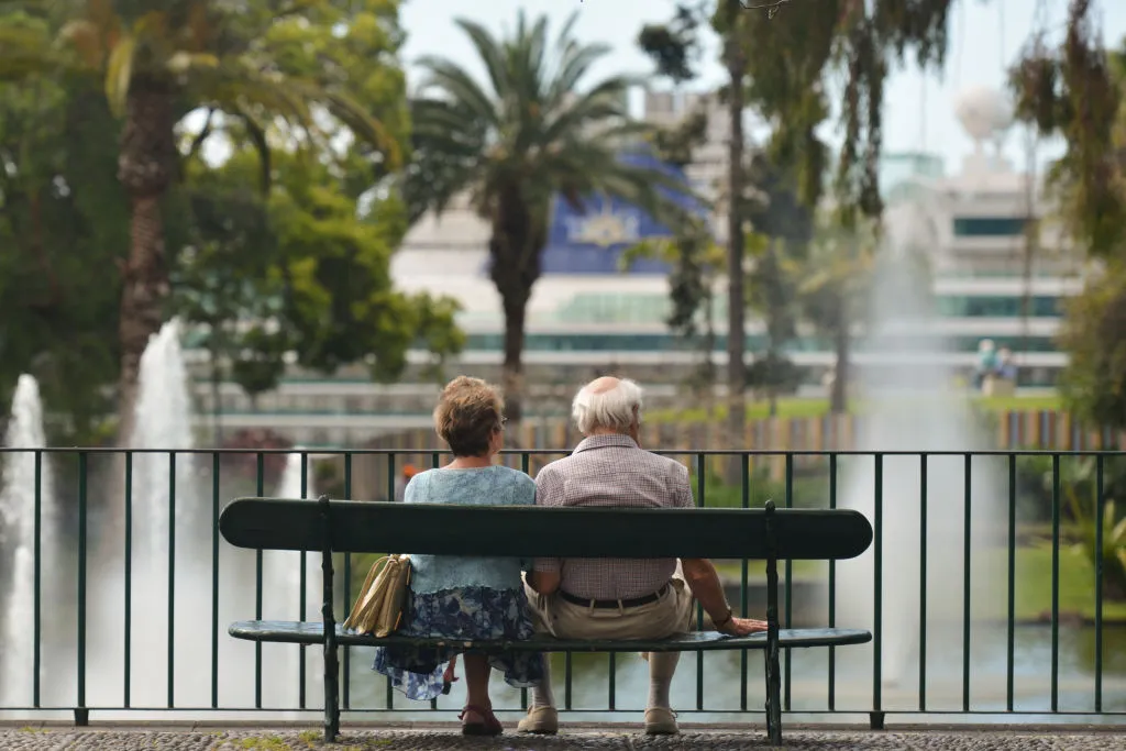 A elderly couple sits on a bench near a lake.