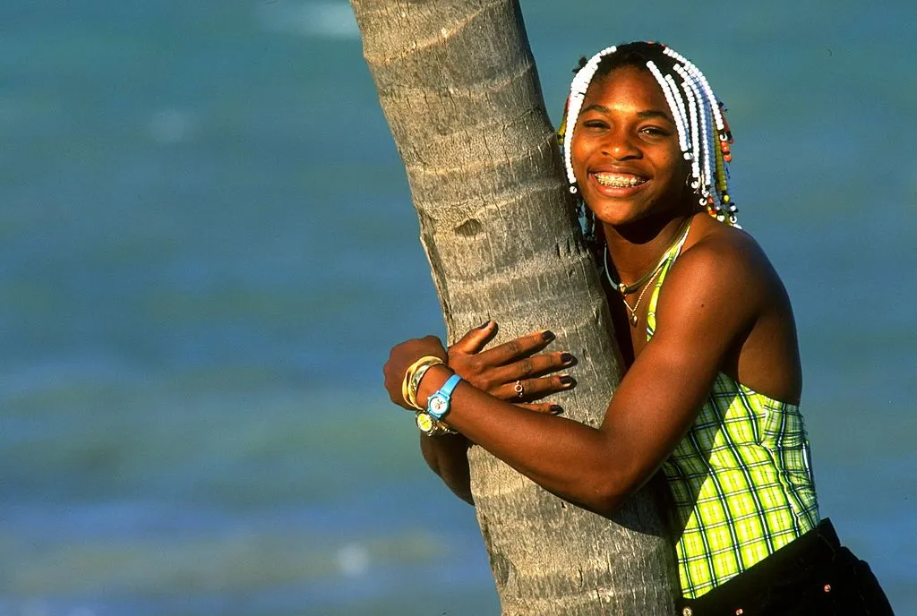Teenage Serena Williams hugs a palm tree and smiles at the camera.