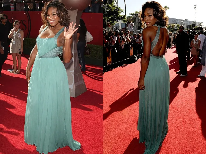 Serena Williams waves to photographers on a red carpet wearing a blue-green gown.
