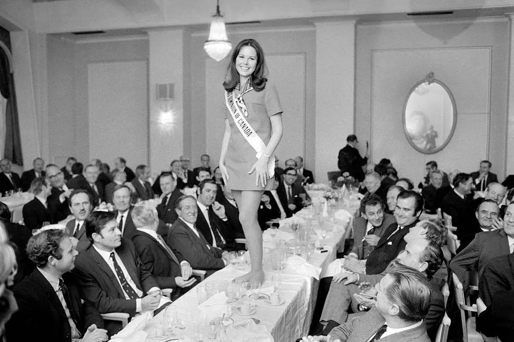 a beauty pageant winner standing barefoot on a table filled with men