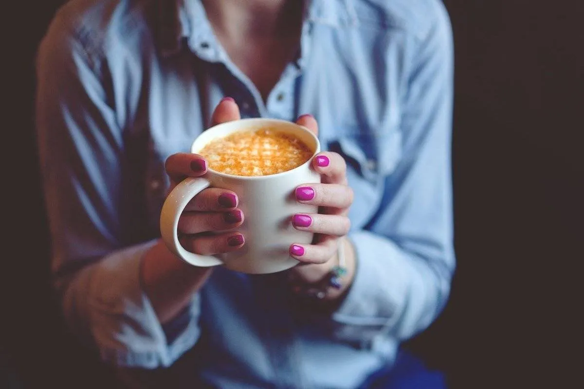 A woman in a blue shirt holds a latte in a mug.