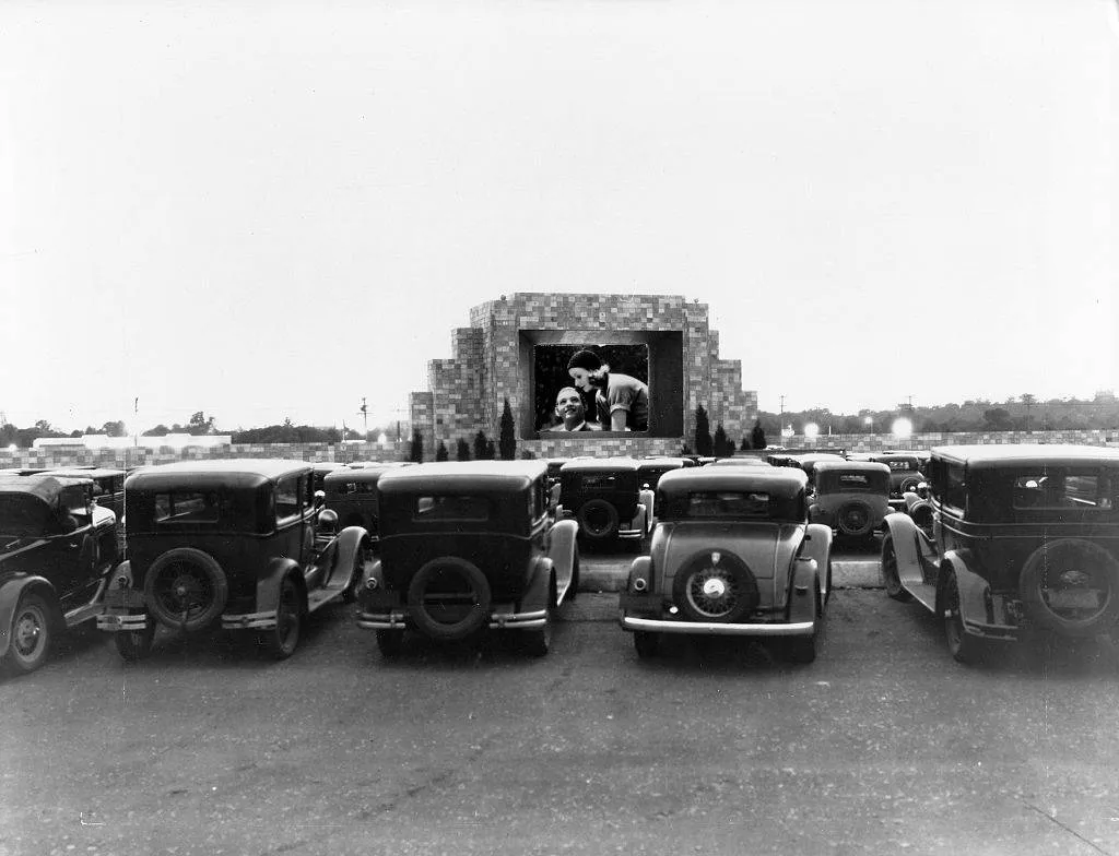 old 30s cars sitting in a drive-in movie theater with a black and white movie on the screen