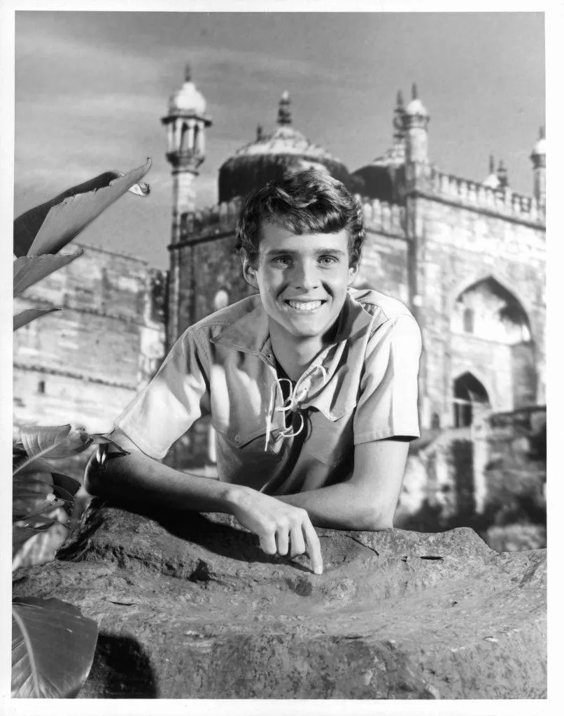 Jay North leaning against a rock with ornate buildings in the background in a scene from the film 'Maya',