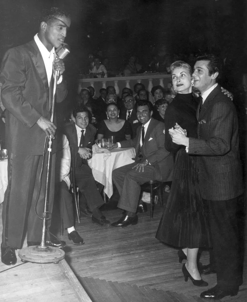 American singer and actor Sammy Davis, Jr. (1925 - 1990) sings on stage at a microphone, as married American actors Janet Leigh and Tony Curtis dance