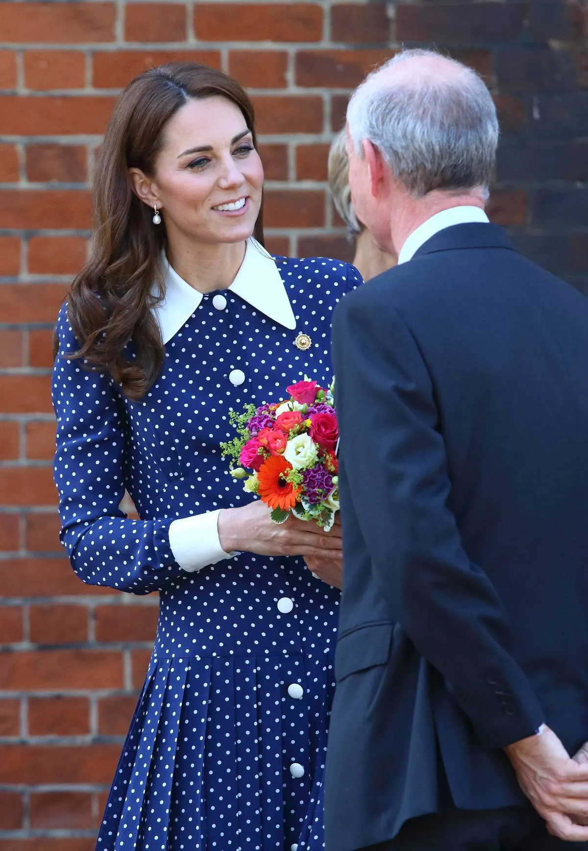 Kate Middleton speaks to a man while wearing a blue polka dot dress with a white collar.