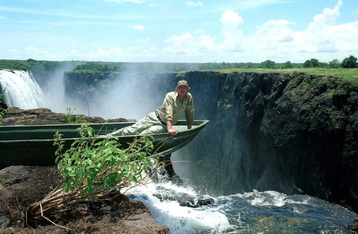 A man sits in a boat that sits in Devil's Pool and leans over Victoria Falls.