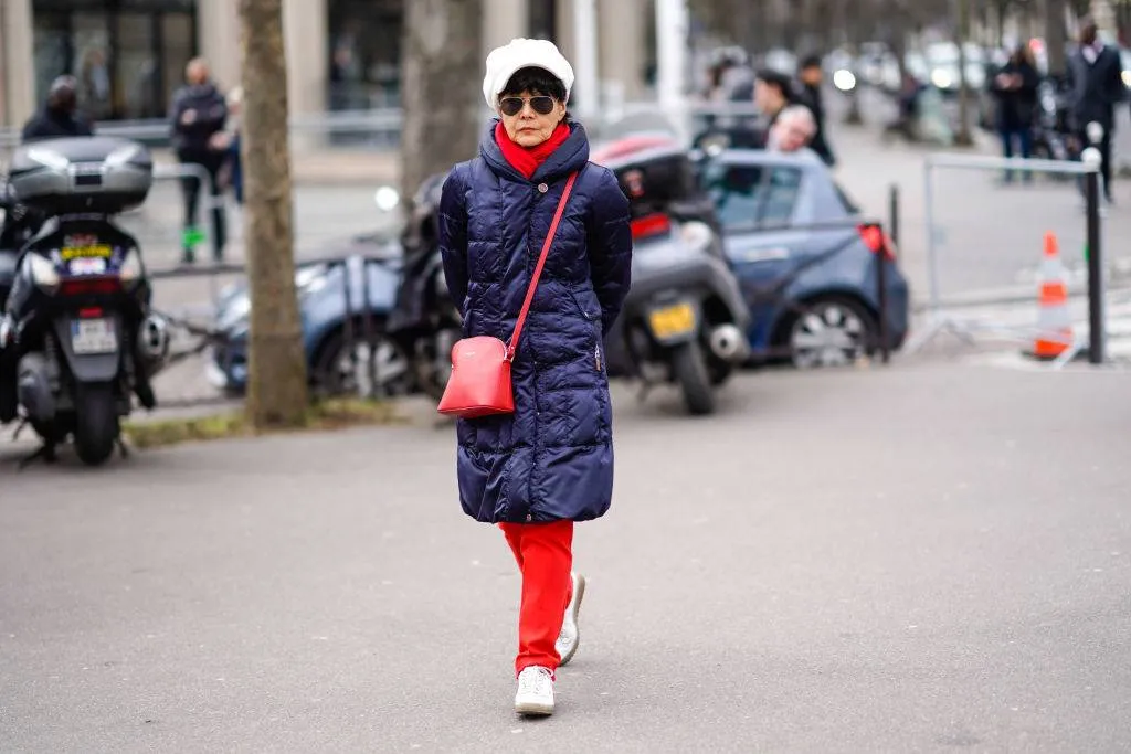 a lady wearing a white beret, sunglasses, a red scarf, a blue long puffer coat, a red bag, red pants, white shoes
