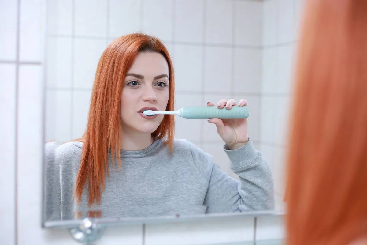 A woman brushes her teeth with an electric toothbrush while looking into a mirror.