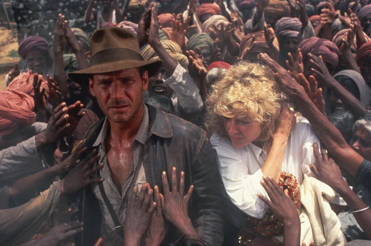 Harrison Ford and Kate Capshaw walking through crowd of beggars as Indiana Jones and Willie Scott